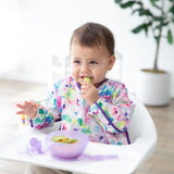 A baby in a floral bib sits in a high chair by a potted plant, nibbling from avocado from a Bumkins Silicone First Feeding Set: Purple Jelly.