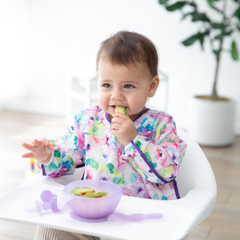 A baby in a floral bib sits in a high chair by a potted plant, nibbling from avocado from a Bumkins Silicone First Feeding Set: Purple Jelly.