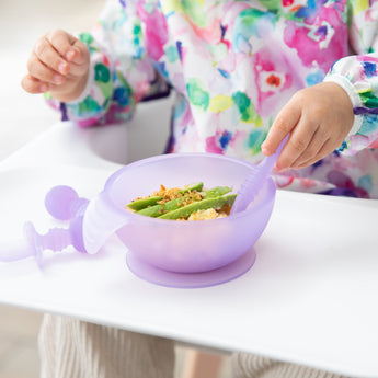 Close-up of a child in a colorful bib eating avocado from a Bumkins Silicone First Feeding Set: Purple Jelly on a high chair tray.