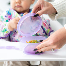 Load image into Gallery viewer, An adult opens a Bumkins Silicone First Feeding Set: Purple Jelly in front of a toddler in a floral bib, seated eagerly in a high chair.
