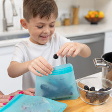 Load image into Gallery viewer, A smiling child in a kitchen places blackberries into a Bumkins Silicone Flat Reusable Bag 2 Pack, Blue, with a bowl nearby.
