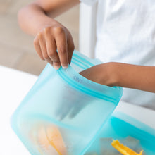 Load image into Gallery viewer, A hand puts orange slices into a Bumkins Silicone Flat Reusable Bag 2 Pack, Blue at a white table.

