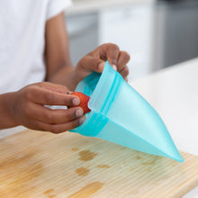 Load image into Gallery viewer, Hands place a strawberry into a Bumkins Silicone Flat Reusable Bag 2 Pack, Blue over a wooden cutting board.
