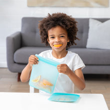 Load image into Gallery viewer, At a table, a child joyfully holds a Bumkins Silicone Flat Reusable Bag 2 Pack, Blue while they smile with an orange slice in their mouth.
