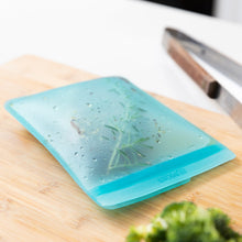 Load image into Gallery viewer, A Bumkins Silicone Flat Reusable Bag 2 Pack, Blue filled with herbs and meat sits on a wood cutting board on a kitchen counter.
