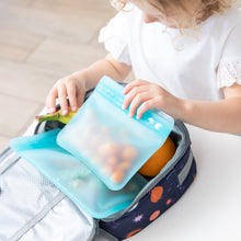 Load image into Gallery viewer, A child packs snacks into an eco-friendly lunchbox at a white table using Bumkins Silicone Flat Reusable Bag 2 Pack, Blue.

