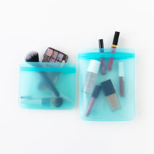 Load image into Gallery viewer, Bumkins Silicone Flat Reusable Bag 2 Pack, Blue holds makeup essentials on a white background—brushes, lipsticks, and an eyeshadow palette.
