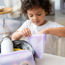 Load image into Gallery viewer, A child places food in a Bumkins Silicone Flat Reusable Bag 2 Pack, Lavender, with a lunchbox and water bottle on the table nearby
