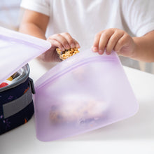 Load image into Gallery viewer, A child's hands place a granola bar in a Bumkins Silicone Flat Reusable Bag 2 Pack, Lavender near a lunchbox on a white table.
