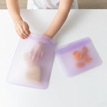 Load image into Gallery viewer, A child places a sandwich in a Bumkins Silicone Flat Reusable Bag 2 Pack, Lavender, with fruit in the other beside it, on a white table.
