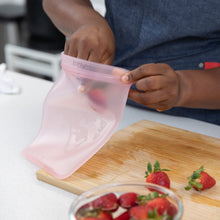 Load image into Gallery viewer, A person places strawberries into a Bumkins Silicone Flat Reusable Bag 2 Pack, Pink on a cutting board with freshly cut strawberries.
