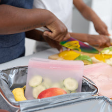Load image into Gallery viewer, Two people chop veggies in a kitchen while a Bumkins Silicone Flat Reusable Bag 2 Pack, Pink with cucumber slices sits in an open lunchbox.
