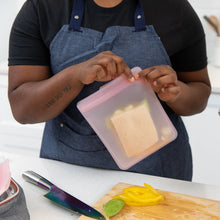 Load image into Gallery viewer, A person in a blue apron holds a sandwich in a Bumkins Silicone Flat Reusable Bag 2 Pack, Pink by sliced bell peppers on a cutting board.
