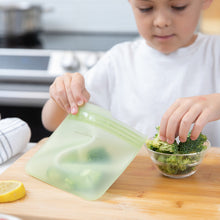 Load image into Gallery viewer, A child puts broccoli in a Bumkins Silicone Flat Reusable Bag 2 Pack, Sage on a kitchen counter.
