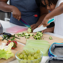Load image into Gallery viewer, Two people chop cucumbers, surrounded by grapes, veggies, and Bumkins Silicone Flat Reusable Bag 2 Pack, Sage nearby on a kitchen counter.
