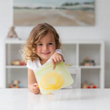 Load image into Gallery viewer, A child smiles at a white table holding a Bumkins Silicone Flat Reusable Bag 2 Pack, Sage, with shelves in the background.
