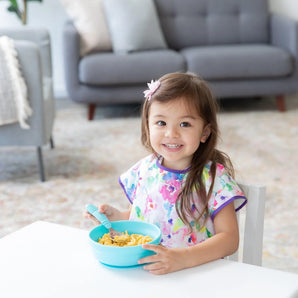 A cheerful girl in a floral bib sits at a white table, eating macaroni & cheese from a Bumkins Silicone Grip Bowl: Blue with a spoon.