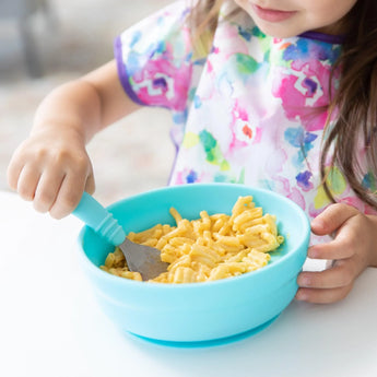 A toddler eats mac and cheese from Bumkins Silicone Grip Bowl: Blue at a white table, while wearing a vibrant bib.