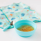 Bumkins Silicone Grip Bowl: Blue with soup and pasta sits in front of a spoon and a blue bib on a white background.