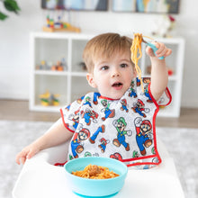 Load image into Gallery viewer, A boy wearing Bumkins Junior Bib: Super Mario™ &amp; Luigi in a high chair looks surprised as he holds spaghetti on a fork from a blue bowl.
