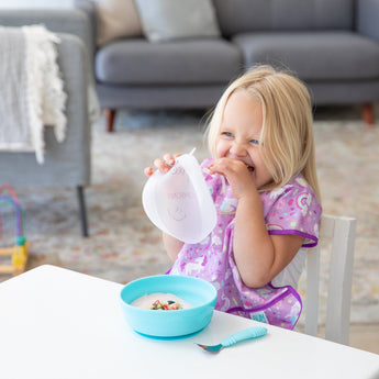 A girl in a purple bib giggles, holding Bumkins Silicone Stretch Lid for Grip Bowl above a blue bowl with yogurt on a white table.