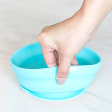 Load image into Gallery viewer, A hand holds a Bumkins Silicone Grip Bowl: Blue, demonstrating the suction cup bottom on a marble surface.
