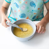 A child in an ocean-themed bib holds a spoon over Bumkins Silicone Grip Bowl: Gray filled with soup and pasta at a white table.