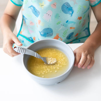 A child in an ocean-themed bib holds a spoon over Bumkins Silicone Grip Bowl: Gray filled with soup and pasta at a white table.