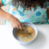 A child enjoys applesauce from Bumkins Silicone Grip Bowl: Gray, spoon in hand, wearing a colorful ocean-themed bib at a white table.