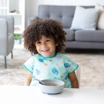 A curly-haired child in a blue bib happily eats from a Bumkins Silicone Grip Bowl: Gray with a spoon at a white table in a bright room.