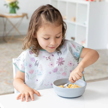 A young girl eats cereal from a Bumkins Silicone Grip Bowl: Gray with a gray spoon, wearing a floral bib at a white table.