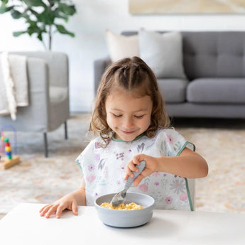 A young girl enjoys cereal from Bumkins Silicone Grip Bowl: Gray with a spoon at a white table, with a cozy living room in the background.