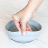 A hand holds a Bumkins Silicone Grip Bowl: Gray, demonstrating the suction cup bottom on a marble surface.