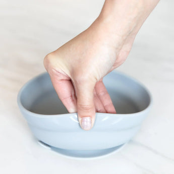 A hand holds a Bumkins Silicone Grip Bowl: Gray, demonstrating the suction cup bottom on a marble surface.