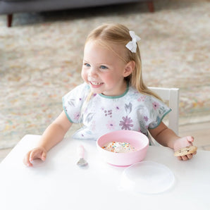 A happy toddler in a floral bib holds a cookie and enjoys ice cream from a Bumkins Silicone Grip Bowl: Pink at a table.