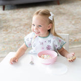 A happy toddler in a floral bib holds a cookie and enjoys ice cream from a Bumkins Silicone Grip Bowl: Pink at a table.
