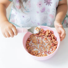 Load image into Gallery viewer, A child wearing a floral bib enjoys colorful cereal and milk from Bumkins Silicone Grip Bowl: Pink at a white table, using a spoon.
