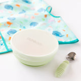 Bumkins Silicone Stretch Lid for Grip Bowl covers a green bowl next to a green spoon and an ocean-themed bib on a white background.