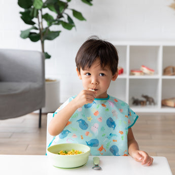A boy wearing a blue whale themed bib looks curiously to the side as he eats from a Bumkins Silicone Grip Bowl: Sage with matching utensils.