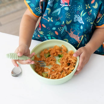 A child in a colorful bib eats pasta from a Bumkins Silicone Grip Bowl: Sage with a fork at a white table.