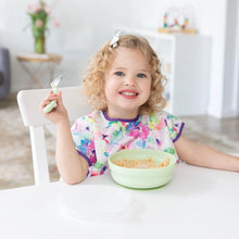 Load image into Gallery viewer, A curly-haired child beams, enjoying cereal from a Bumkins Silicone Grip Bowl: Sage, adding charm with their bright bib and bow.
