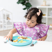 Load image into Gallery viewer, A girl wearing a bow and Bumkins Ruffle Apron Bib: Unicorns picks up fruit with her fingers from a blue plate with blue utensils nearby.
