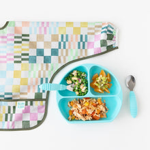 Load image into Gallery viewer, Bumkins Sleeved Bib: Motley Check lays flat next to a blue divided plate with fruit &amp; veggies and blue utensils on a white background.
