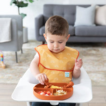 Load image into Gallery viewer, A toddler enjoys a meal from Bumkins Silicone Grip Dish: Clay in a high chair, with a cozy gray sofa in the background.
