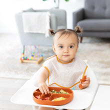 Load image into Gallery viewer, A toddler with pigtails in a high chair enjoys a meal from Bumkins Silicone Grip Dish: Clay, wearing a patterned bib in a cozy living room.
