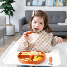 Load image into Gallery viewer, A smiling girl in a tan checkered bib uses Bumkins Spoon + Fork: Clay to eat from a matching plate on a high chair tray.
