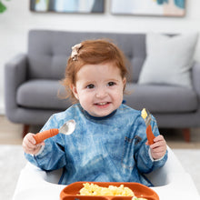 Load image into Gallery viewer, A red-haired girl wearing Bumkins Sleeved Bib: Vintage Denim smiles, holding orange utensils over a plate of eggs in front of a gray couch.
