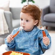 Load image into Gallery viewer, A red-haired girl wearing a blue bib places Bumkins Spoon + Fork: Clay in her mouth as she eats eggs in a cozy living room.
