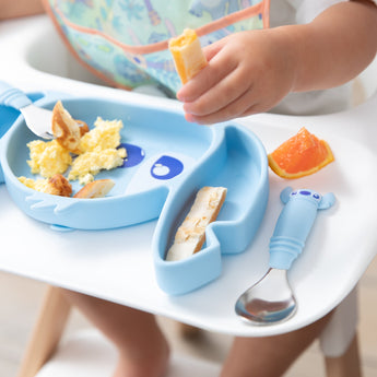 Close-up of a child in a high chair holding food over a Bumkins Silicone Grip Dish: Stitch with a matching spoon on the tray.