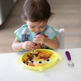 A toddler self-feeds from a Bumkins Silicone Grip Dish: Toy Story Alien with purple utensils at a white table.